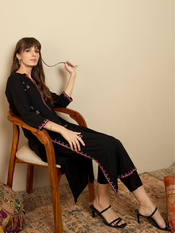 Woman sitting on a chair wearing a black outfit with pink embroidery against a beige wall.